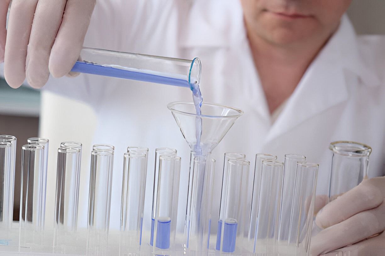 A laboratory technician in a white coat and gloves pouring a specimen through a funnel into a rack of glass test tubes