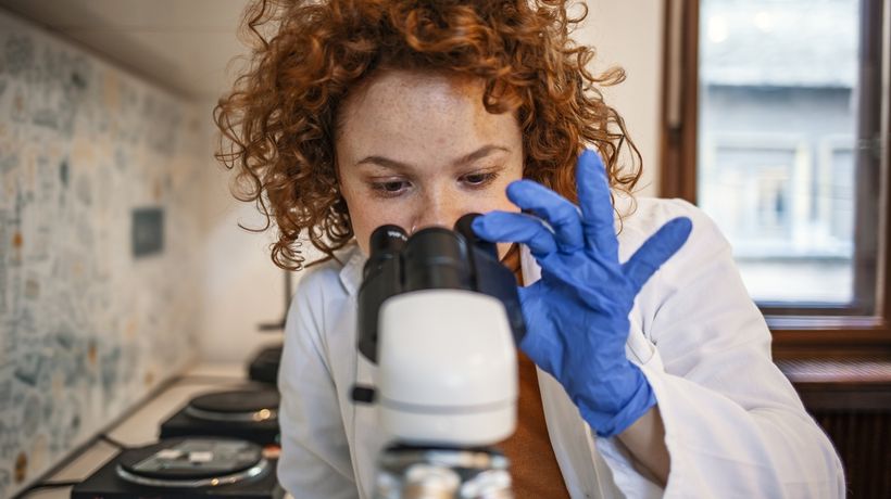 A laboratory scientist in a white coat and gloves looking through a microscope
