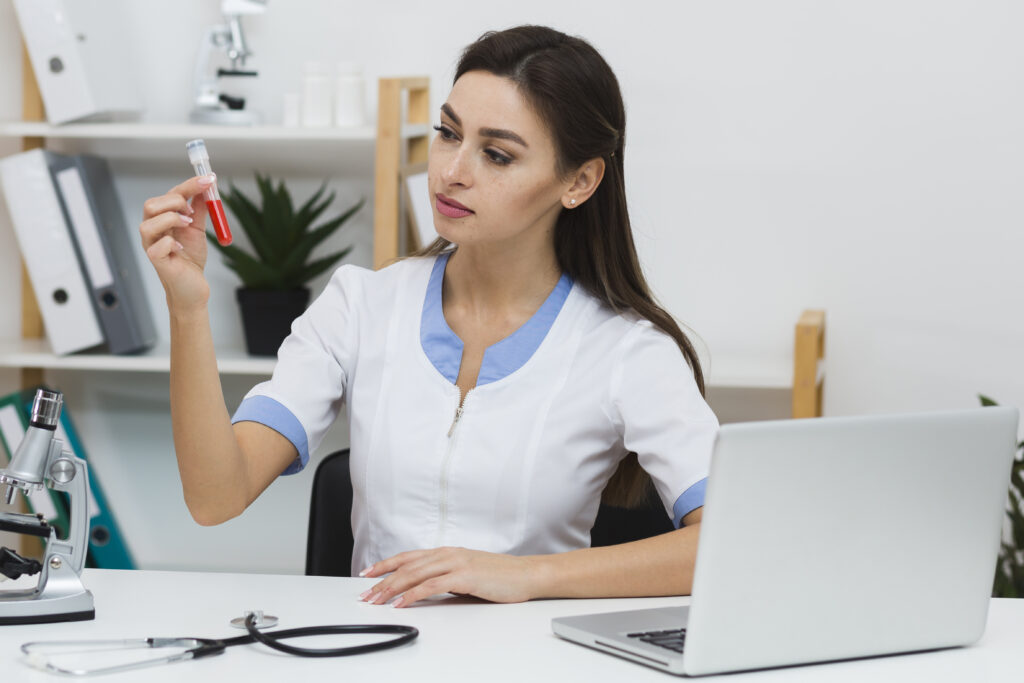 A clinician in a lab coat holding a blood collection tube up to the light at a desk, with a microscope and stethoscope beside her